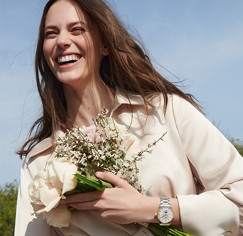 A female model wearing a Longines watch and a cream coloured shirt with a blue sky background.