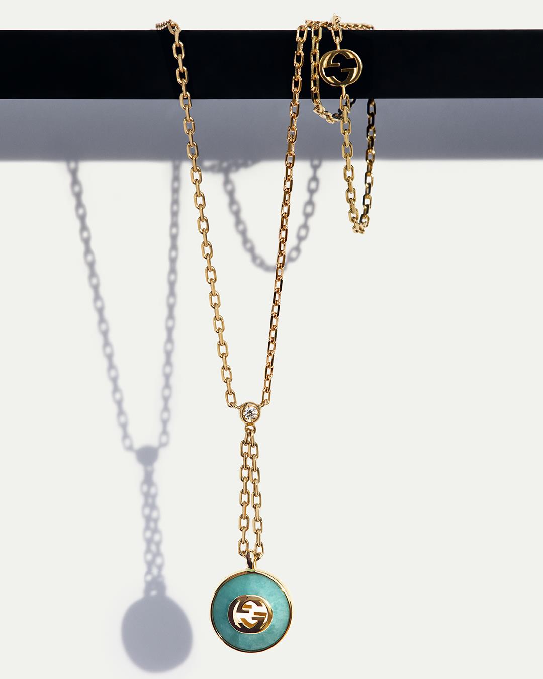 A yellow gold necklace with a green Gucci Interlocking emblem hanging against an off-white background.