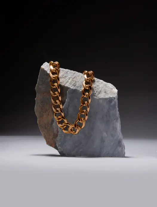A yellow gold, large link chain laying on a grey coloured rock on a black background.